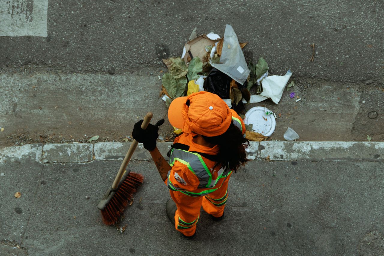 services-02 Vue plongeante d’un agent de propreté en uniforme orange balayant des débris sur un trottoir de la ville.