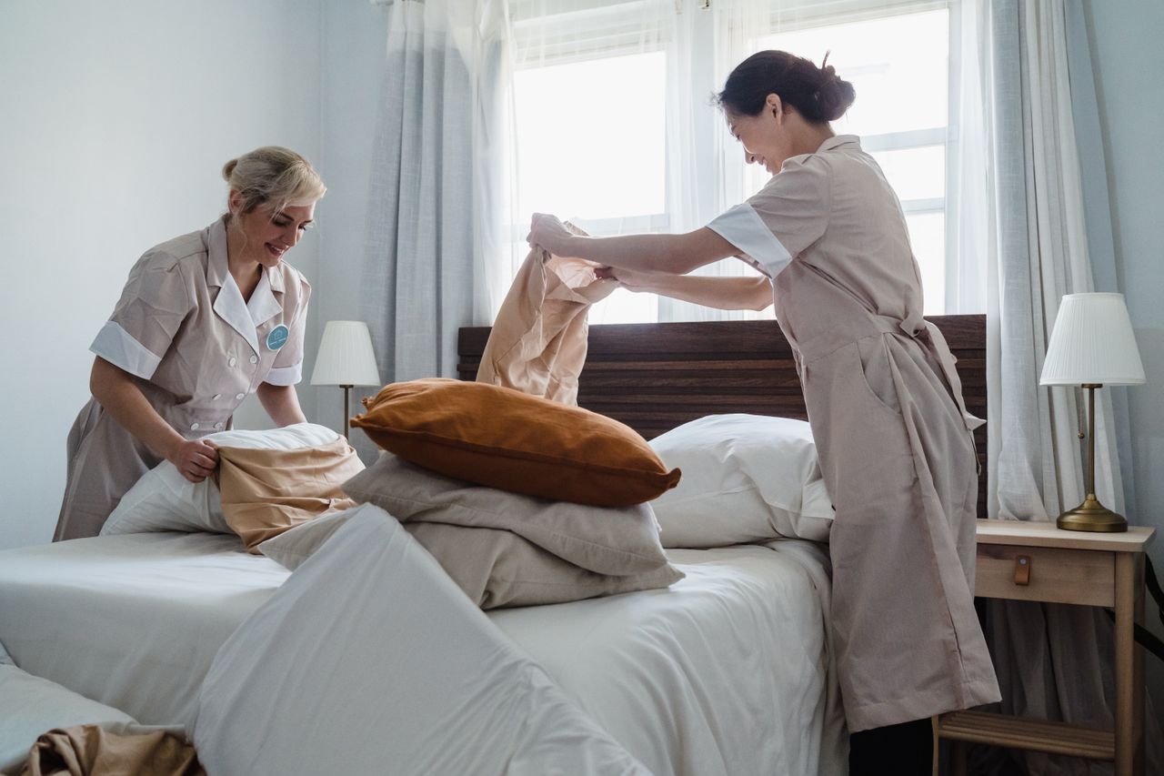 Two housekeepers in uniform are making up a hotel room bed with fresh linen and pillows.